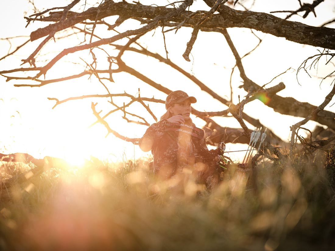 A woman sits under a tree near sunset with a PSE bow.